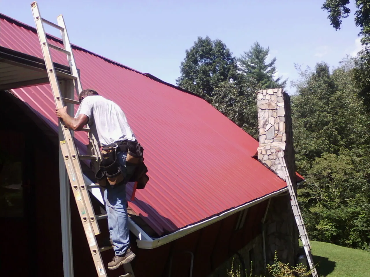 Roofer on a ladder beside a red metal roof and stone chimney — Soffit & Fascia Repair by Roofing Brothers in Moreland